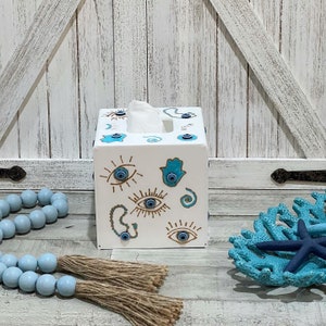 May include: White wooden tissue box cover with a blue and gold design featuring multiple evil eye symbols. The box is sitting on a wooden surface with blue beads and a blue starfish.