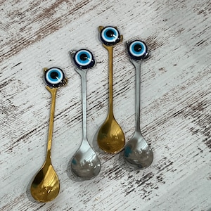 May include: Four silver spoons with blue and white evil eye charms on the handles. The spoons are on a white wooden surface.