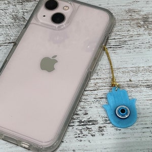 May include: A pink iPhone case with a clear back and a gold chain attached to a blue hamsa hand charm with an evil eye design.