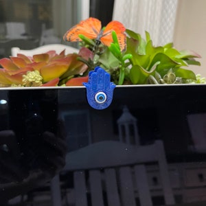 May include: A blue Hamsa hand webcam cover with a central blue eye design. The cover is placed on a laptop, with a decorative arrangement of succulents and an orange butterfly in the background.