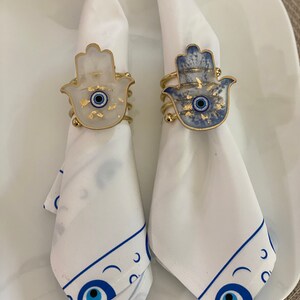 May include: Two white napkins with blue and gold hamsa hand napkin rings. The rings feature a blue evil eye in the centre of the hamsa hand.