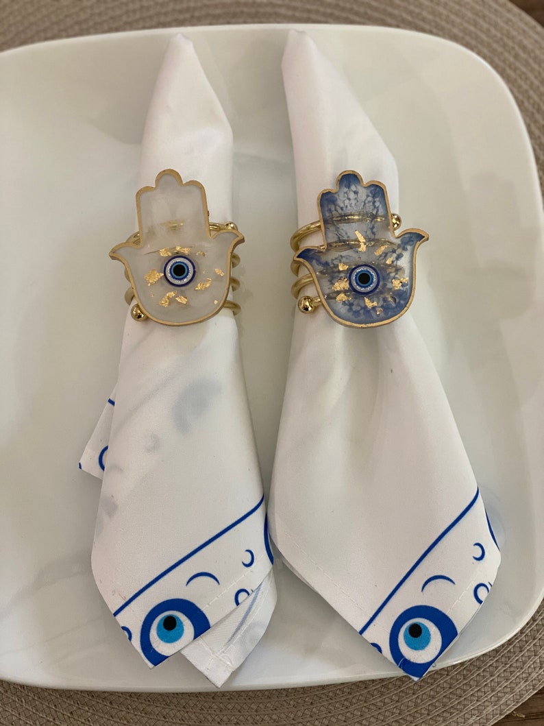 May include: Two white napkins with blue and gold hamsa hand napkin rings. The rings feature a blue evil eye in the centre of the hamsa hand.