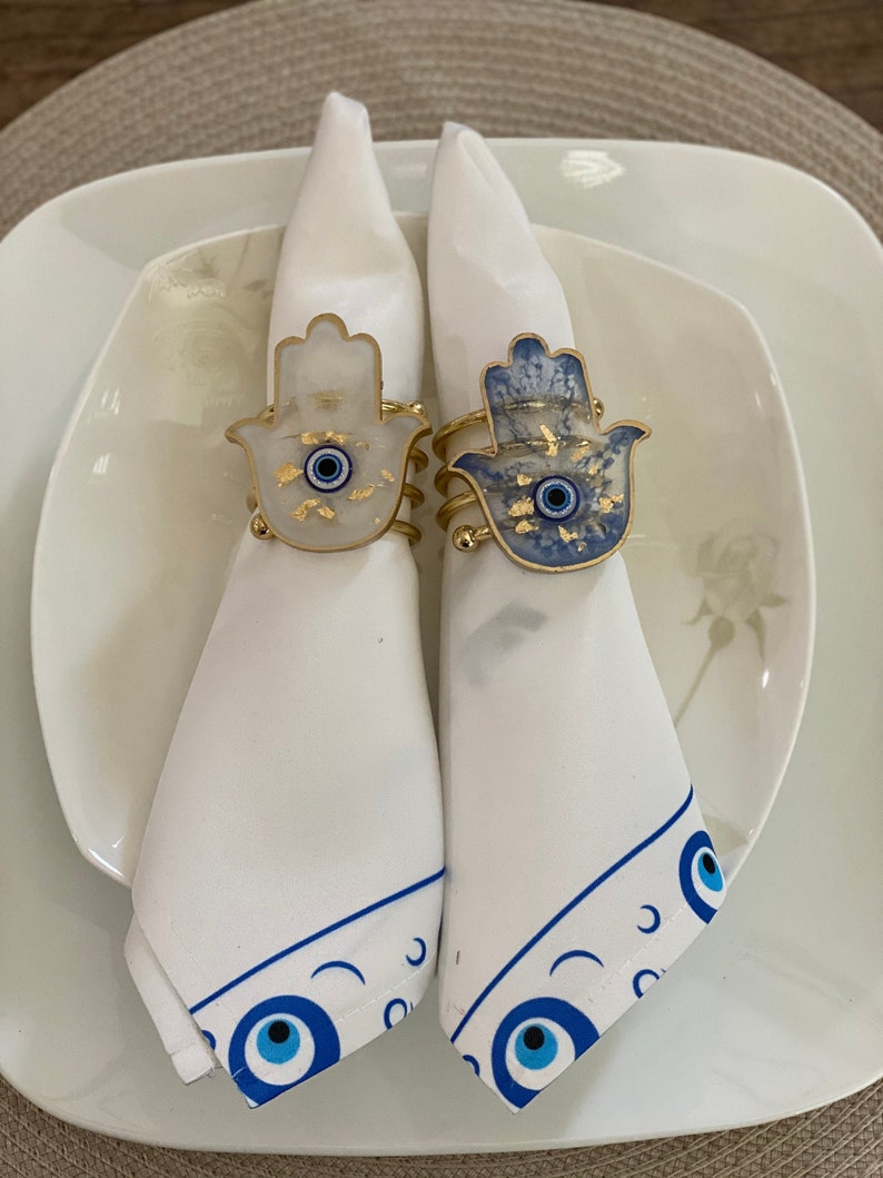 May include: Two white napkins with blue and gold hamsa hand napkin rings. The rings feature a blue evil eye in the centre of the hamsa hand.