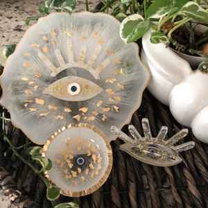 May include: Three white resin trays with gold accents and blue evil eye designs. The trays are in various sizes and shapes, and are perfect for displaying small items or jewellery.