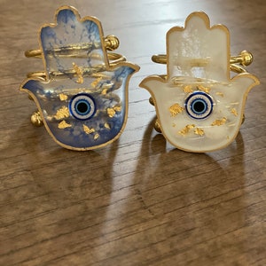 May include: Two gold hamsa hand napkin rings with blue evil eye charms. One ring has a white background with gold flecks and the other has a blue background with gold flecks.