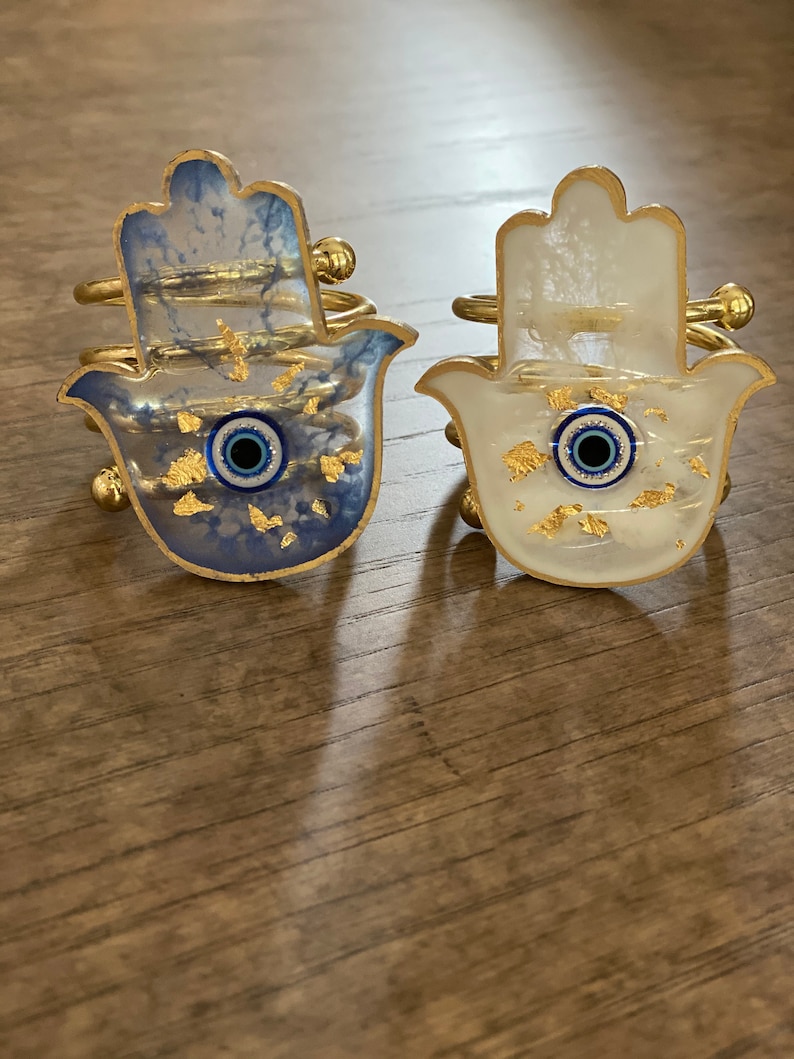 May include: Two gold hamsa hand napkin rings with blue evil eye charms. One ring has a white background with gold flecks and the other has a blue background with gold flecks.