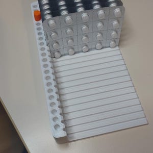 May include: A stack of silver blister packs containing white pills rests on a white, ribbed tray with a side rail. An orange cylindrical piece is visible on the side rail. The pills are likely medication or supplements.
