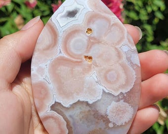 May include: A teardrop-shaped pink and white stone with a unique, swirling pattern. The polished surface reveals concentric circles and natural variations in color, creating a marbled effect. The stone is held in a hand, showcasing its size and texture.