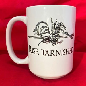 May include: White ceramic mug featuring a black and white illustration of a sword and flowers. The text "Rise, Tarnished" is printed on the mug.