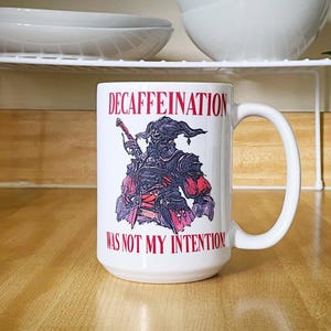 Puede incluir: Taza de cerámica blanca con el texto rojo «DECAFFEINATION WAS NOT MY INTENTION». La taza presenta una ilustración detallada de un personaje de fantasía en negro, rojo y morado. La taza tiene un asa grande.