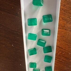 Op de afbeelding: Een verzameling smaragdgroene edelstenen in een witte doos. De edelstenen zijn rechthoekig en hebben een gepolijste afwerking.