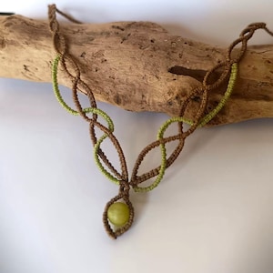 Può includere: Collana in macramè fatta a mano con un design a cordoncino marrone e dettagli verde oliva. La collana presenta un pendente centrale con una pietra rotonda verde chiaro. La collana è esposta su un pezzo di legno.
