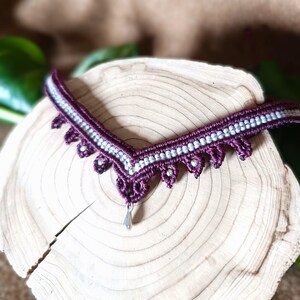 Peut inclure: Un collier en macramé fait à la main en forme de V, avec un design violet foncé et blanc. Le collier présente un motif délicat et dentelé avec de petits accents pendants et un pendentif en forme de goutte d'eau argenté. Il est présenté sur une surface en bois.