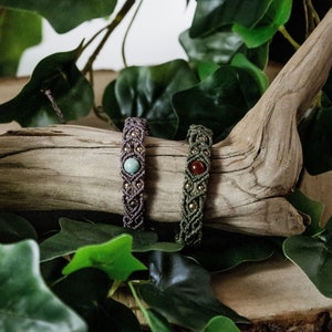 Pode incluir: Duas pulseiras de macramé feitas à mão com contas de pedras preciosas. Uma pulseira é marrom com uma pedra verde e a outra é verde com uma pedra vermelha. As pulseiras são exibidas em um pedaço de madeira à deriva.