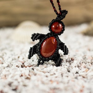Peut inclure: Un pendentif en macramé noir en forme de tortue avec une pierre de cornaline rouge. Le pendentif est sur un fond blanc.