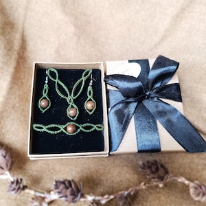 Peut inclure: Un ensemble de bijoux faits à la main comprenant un collier, des boucles d'oreilles et un bracelet en macramé vert avec des perles en bois. L'ensemble est présenté dans une petite boîte avec un intérieur en velours noir, à côté d'un coffret cadeau noué d'un ruban de satin noir.