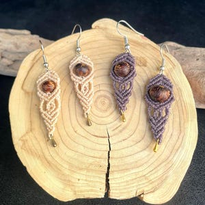 Peut inclure: Quatre boucles d'oreilles en macramé beige et violet, chacune avec un centre en perle de bois et des accents dorés. Les boucles d'oreilles sont présentées sur une tranche de bois, mettant en valeur les nœuds complexes et les matériaux naturels. La longueur des boucles est d'environ 5 cm.