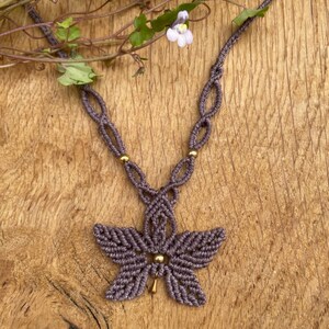 May include: A handmade macrame necklace in a muted purple hue, featuring a flower-shaped pendant with a gold bead center. The necklace is displayed on a wooden surface, with delicate white flowers and green leaves in the background. The necklace's length is approximately 18 inches.