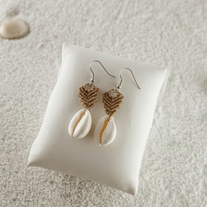 May include: A pair of earrings featuring a brown macrame heart charm and a white seashell pendant. The earrings are presented on a white cushion against a backdrop of white sand.
