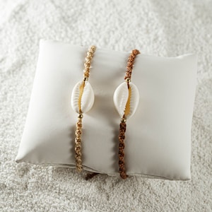 Op de afbeelding: Twee armbanden met kauri schelpen en gouden kralen. De ene armband is gemaakt met bruin koord en de andere met beige koord. De armbanden worden getoond op een wit kussen.