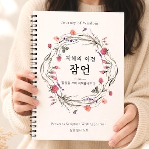 May include: A spiral-bound journal with the text "Journey of Wisdom" and Korean characters. The cover features a floral wreath design with pink and red flowers. The journal is held by a person wearing a cream-colored sweater.