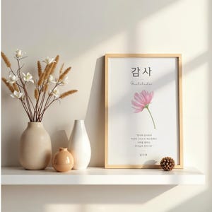 Pode incluir: Uma impressão emoldurada com a palavra coreana "감사" e a palavra inglesa "Gratitude" acima de uma ilustração de flor rosa. A impressão está em uma prateleira branca com vasos e elementos decorativos.