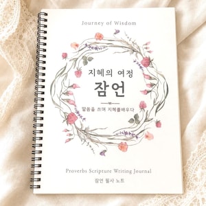 May include: A spiral-bound journal with the title "Journey of Wisdom" and text in Korean. The cover features a floral wreath with pink and orange flowers. Below the title is the text "Proverbs Scripture Writing Journal".