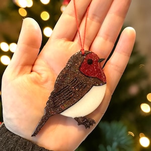 May include: A robin-shaped hanging ornament featuring a dark brown glittery back and wing, a bright red glittery chest, and a solid white belly. The bird has a small black eye and a black beak, and it is suspended by a thin red string for display.