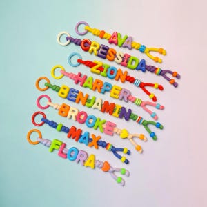 May include: A collection of eight personalised keychains featuring colourful plastic letter beads spelling the names AVA, CRESSIDA, ZION, HARPER, BENJAMIN, BROOKE, MAX, and FLORA. Each keychain includes a circular ring and decorative patterned beads.