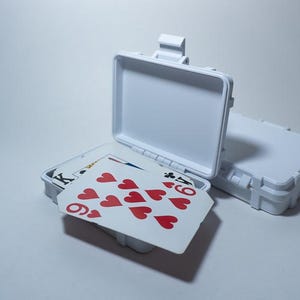 May include: A white plastic case with a lid that opens to reveal a deck of playing cards. The cards are arranged in a fan shape, showing the nine of hearts.