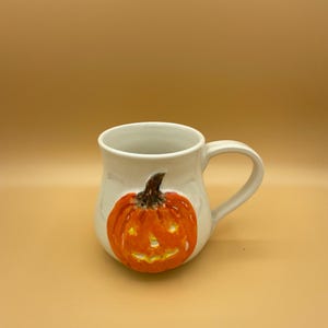 May include: White ceramic mug featuring a painted orange pumpkin design. The pumpkin has a carved face and a brown stem. The mug has a curved handle and is set against a light orange background.