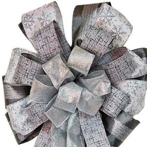 May include: A large, silver and gray bow with a snowflake pattern. The bow is made of multiple layers of ribbon and is tied in a knot in the center.