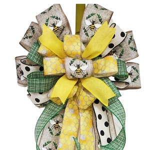 May include: A yellow, green, and brown bee-themed bow with a burlap ribbon and black polka dots. The bow is tied with a yellow ribbon and has a bee design on it.