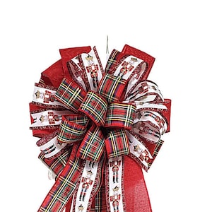 May include: A red and green plaid ribbon bow with a white ribbon with a nutcracker pattern. The bow is tied with a red ribbon and has a wire loop for hanging.