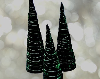 May include: Three dark green velvet cone-shaped decorative trees. Each tree is wrapped with a silver-colored, spiraling ribbon. The trees vary in size, with the tallest being approximately 12 inches high. The background is a soft, blurred bokeh effect.
