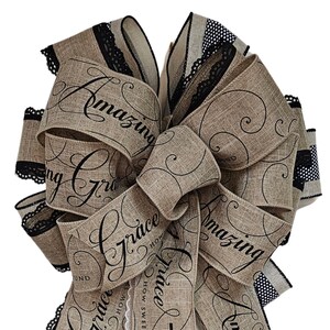 May include: A decorative bow made of tan burlap ribbon featuring black script text that reads Amazing Grace How Sweet the Sound. The ribbon edges are trimmed with black lace and black and white polka dot fabric, creating a layered, full appearance.