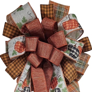 May include: A decorative bow made of multiple ribbons featuring a mix of solid copper-colored fabric, brown and black gingham check, and a light gray ribbon printed with orange and black plaid pumpkins and green leaves on a white background.