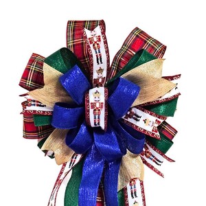 May include: A Christmas bow with a red, green, gold, and blue plaid ribbon. The bow is decorated with a white ribbon with a repeating pattern of nutcracker figures.