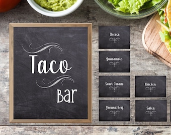 May include: A taco bar setup with a chalkboard sign that reads "Taco Bar". Includes bowls of guacamole and lettuce, ingredient labels, and filled tacos on a wooden board. The scene is set on a rustic wooden table.