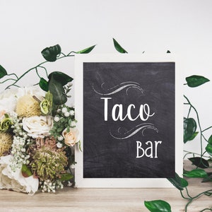 May include: A chalkboard sign with the words "Taco Bar" in white script, framed in white. A bouquet of flowers and greenery are on the left, with vines and leaves around the sign. The sign is on a wooden surface.