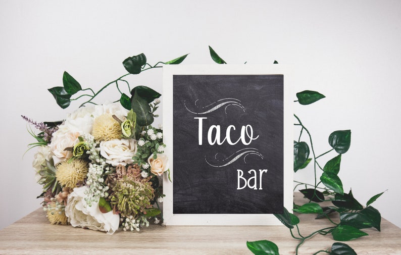 May include: A chalkboard sign with the words "Taco Bar" in white script, framed in white. A bouquet of flowers and greenery are on the left, with vines and leaves around the sign. The sign is on a wooden surface.