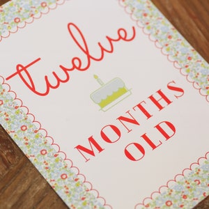 May include: A white card with a red scalloped border and a floral pattern. The card says "twelve months old" in red text with a small illustration of a birthday cake with a single candle.