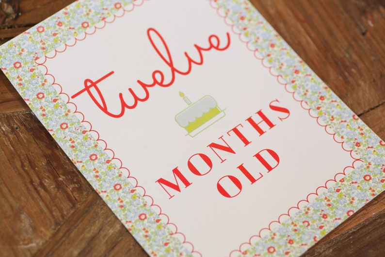 May include: A white card with a red scalloped border and a floral pattern. The card says "twelve months old" in red text with a small illustration of a birthday cake with a single candle.