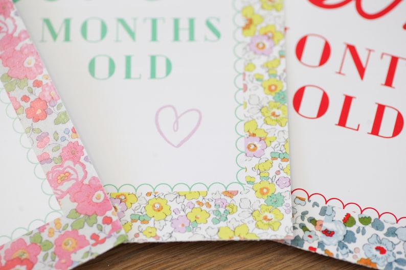 May include: Three white cards with scalloped edges and floral patterned borders. The cards have the text "6 months old", "12 months old", and "24 months old" printed in green, red, and red respectively.