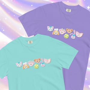 Könnte beinhalten: Zwei T-Shirts mit einer Reihe von Comic-Tiergesichtern, die auf der Vorderseite aufgedruckt sind. Das obere Shirt ist lavendelfarben und das untere Shirt ist türkis.
