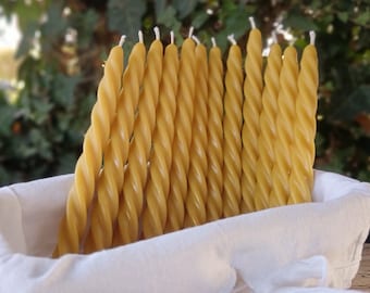 May include: A collection of twelve yellow beeswax taper candles with a twisted, spiral design. Each candle features a smooth, cylindrical shape with a visible white cotton wick at the top. The candles are arranged upright in a white fabric basket.