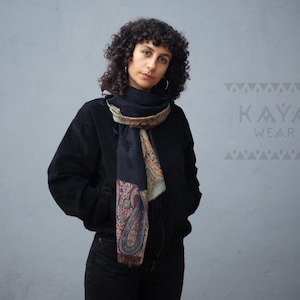 Puede incluir: Una persona con una chaqueta negra y pantalones negros, con una bufanda estampada alrededor del cuello. La bufanda presenta un diseño de cachemira en tonos rojos, dorados y azules. El texto "KAYA WEAR" es visible en la pared.