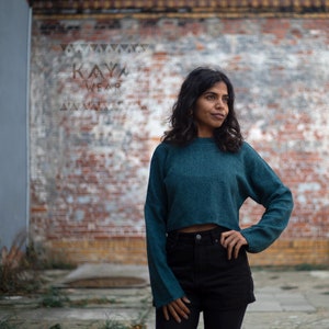 May include: A woman wearing a teal blue cropped sweater with long sleeves and black shorts. She is standing in front of a brick wall with the text "KAYA WEAR" on it.