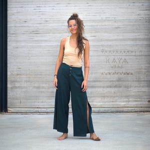 Puede incluir: Una mujer con una camiseta sin mangas beige y pantalones de pierna ancha verde oscuro con botones y aberturas laterales. Los pantalones se combinan con una camiseta sin mangas beige. El fondo presenta una pared de color claro y una puerta de garaje con el texto "KAYA Wear".