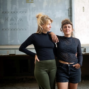 Puede incluir: Dos personas de pie frente a una puerta de metal gris. La persona de la izquierda lleva una camisa negra de manga larga y pantalones verdes. La persona de la derecha lleva una camisa negra de manga larga con una superposición transparente y pantalones cortos de mezclilla negros. El texto "KAYA WEAR" es visible en la puerta.